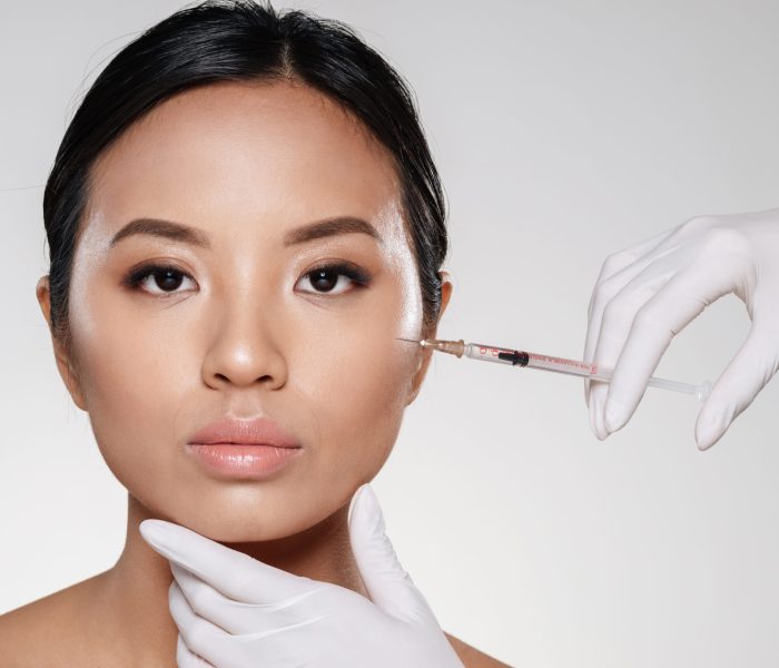 Portrait of a beautiful asian woman getting a beauty medical treatment with a syringe injection isolated over gray background