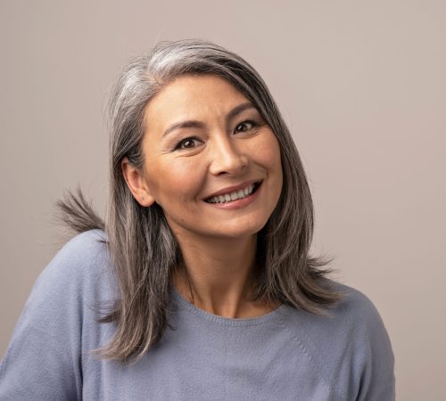 Attractive Senior Asian Woman Broadly Smiles At Camera. Beautiful Grey-Haired Woman Lifts Her Right Shoulder And Sheepishly Smiles. Portrait. Studio Photoshoot